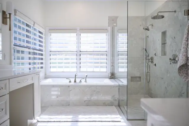 Ballwin primary bathroom by Aptitude Design & Build with a frameless glass shower, marble-look tile and a soaking tub under a window